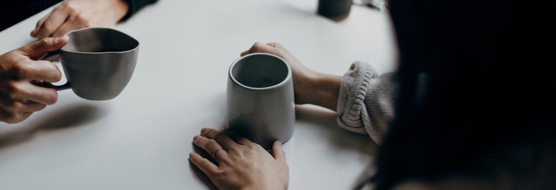 Die Hände zweier Menschen, die am Tisch sitzen und eine Kaffeetasse halten